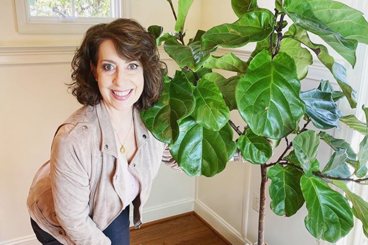 Smiling woman standing next to a large Fiddle Leaf Fig plant in a sunlit room, showcasing the use of plants in interior design.