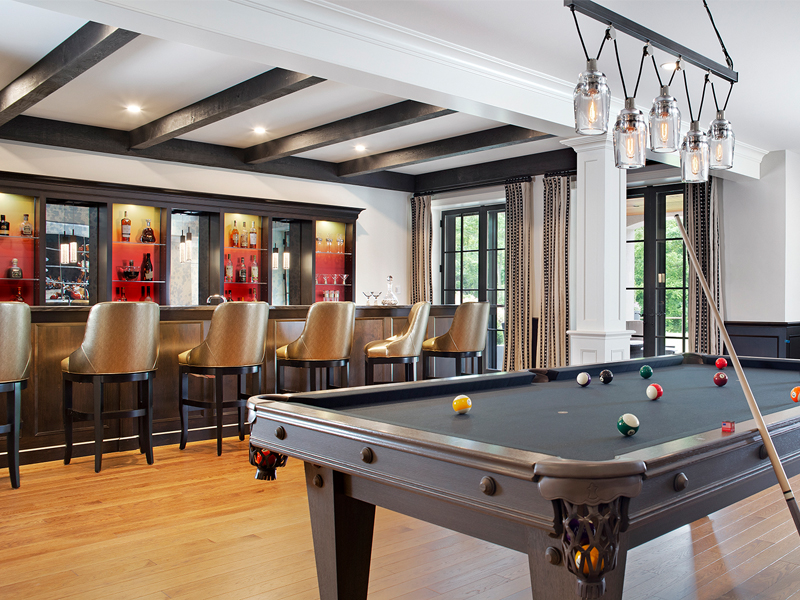 Modern game room with black pool table, sleek home bar, and warm wood floors accented by exposed ceiling beams.